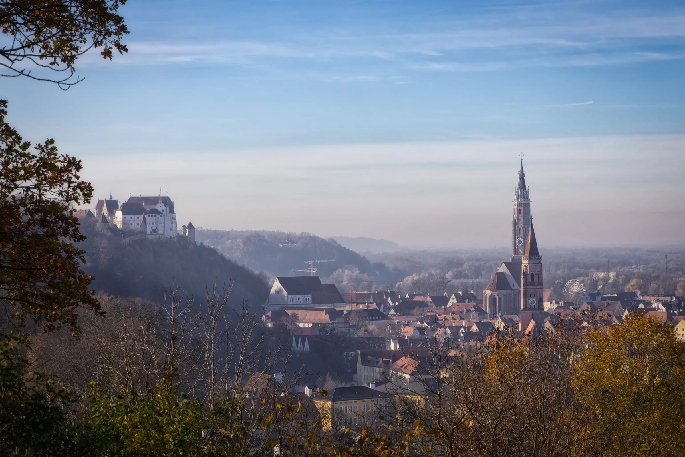 Durch Herbstlaub gerahmter Blick auf Landshut, links Burg Trausnitz auf dem Hügel, rechts der Turm von St. Martin über der Altstadt im Dunst.
