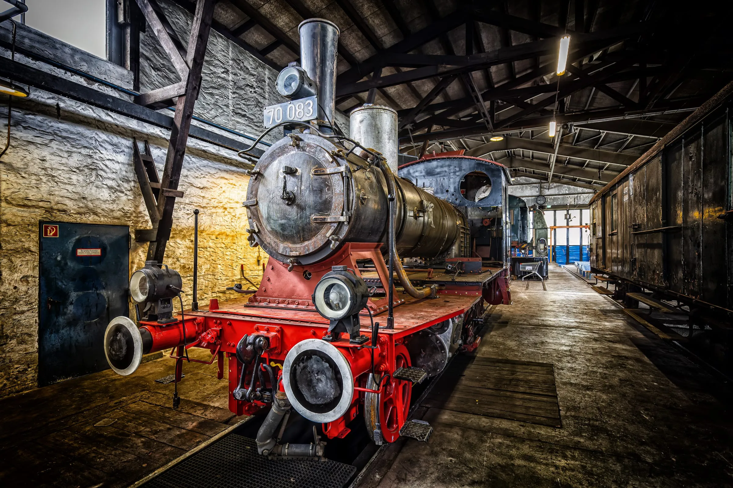 Dampflok 70 083 mit rotem Rahmen und dunklem Kessel in einem Lokschuppen mit dunklem Dachstuhl und Steinwand, rechts ein alter Waggon.
