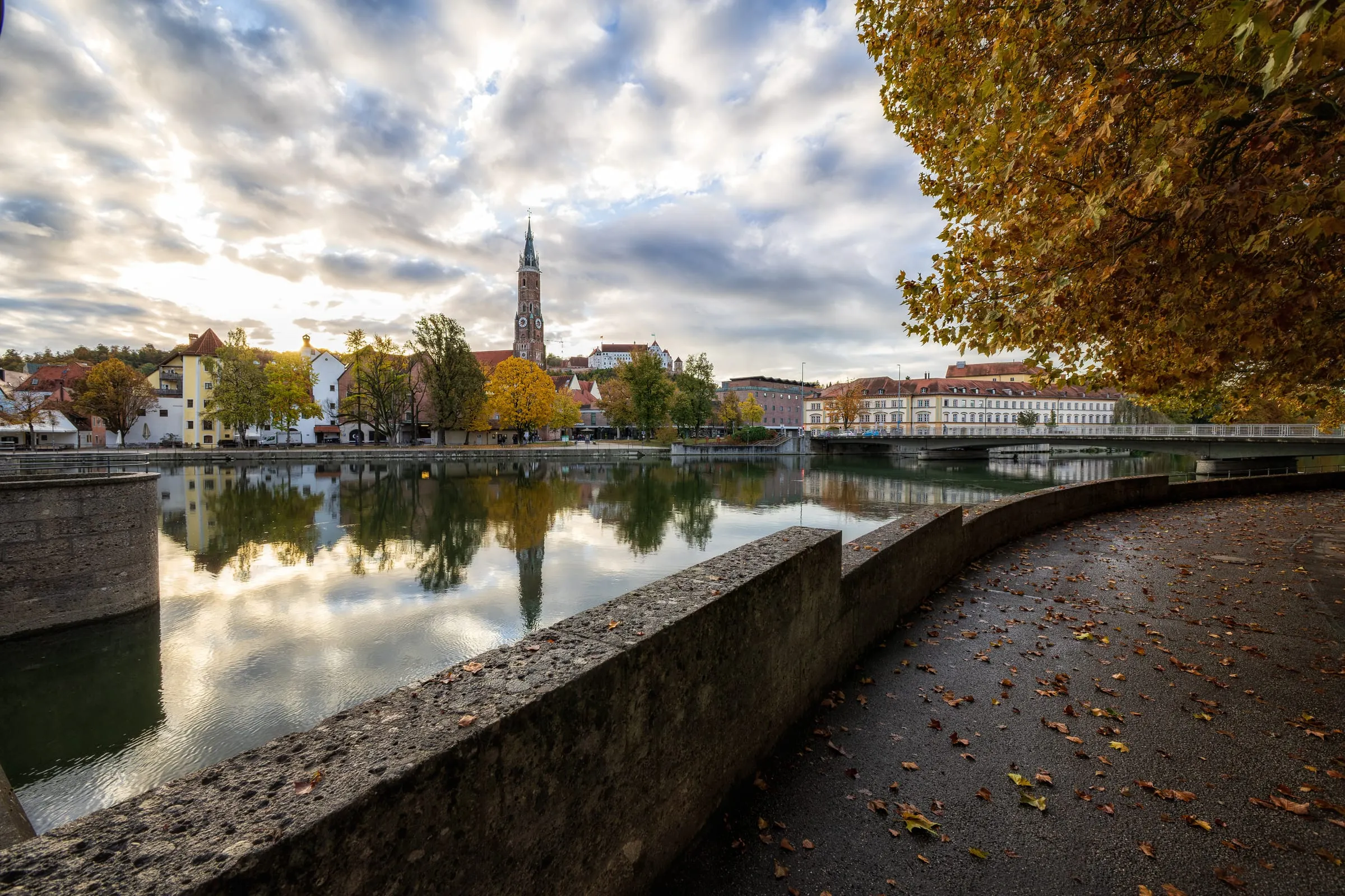 Gebogene Ufermauer und laubbedeckter Weg an der Isar, im Wasser spiegeln sich der Turm von St. Martin und Bäume, rechts eine Brücke unter einem gelben Herbstbaum.