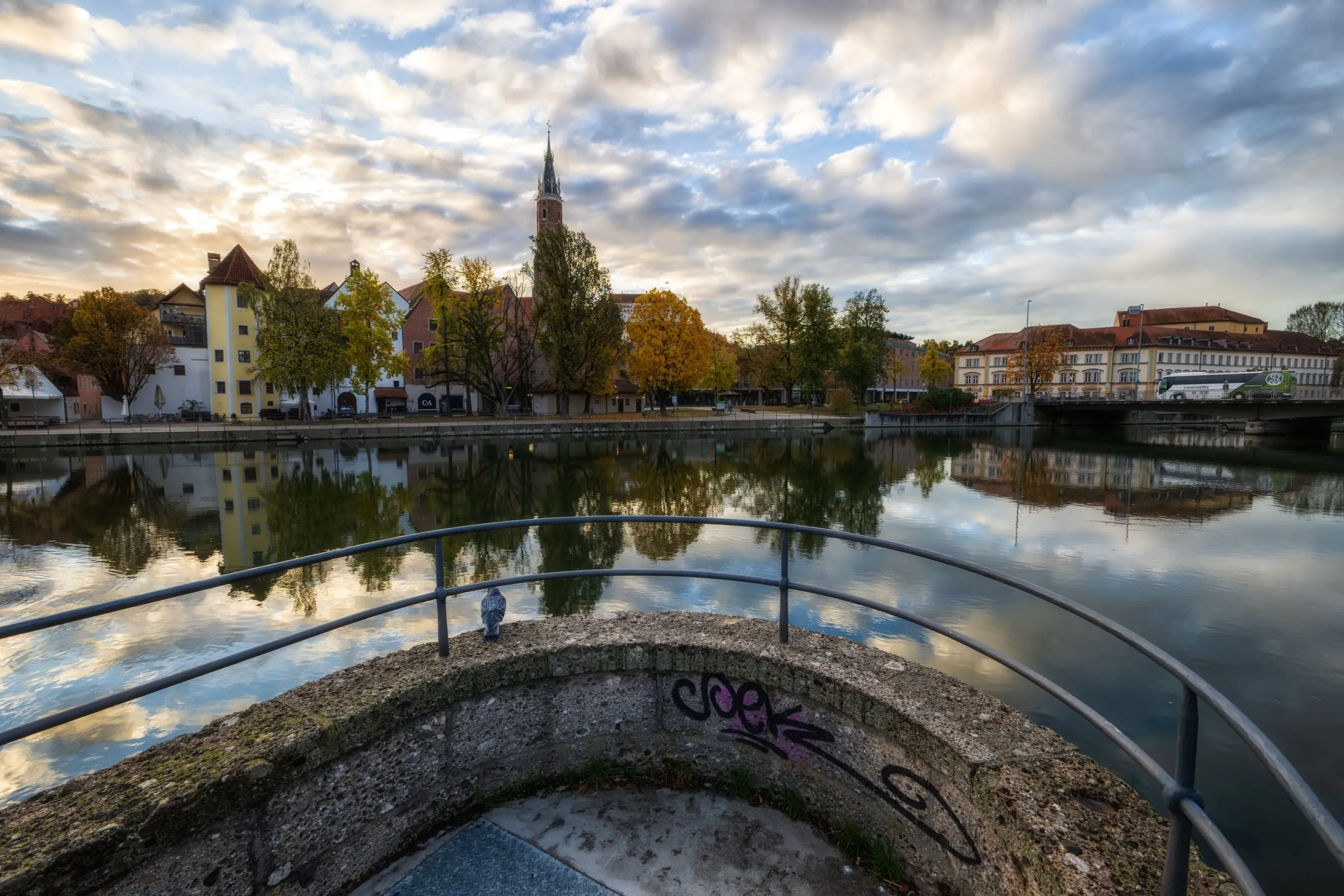 Runde Uferplattform mit Geländer und einer Taube; gegenüber spiegeln sich Häuser und Herbstbäume in der Isar, dahinter der Turm von St. Martin unter Wolken.