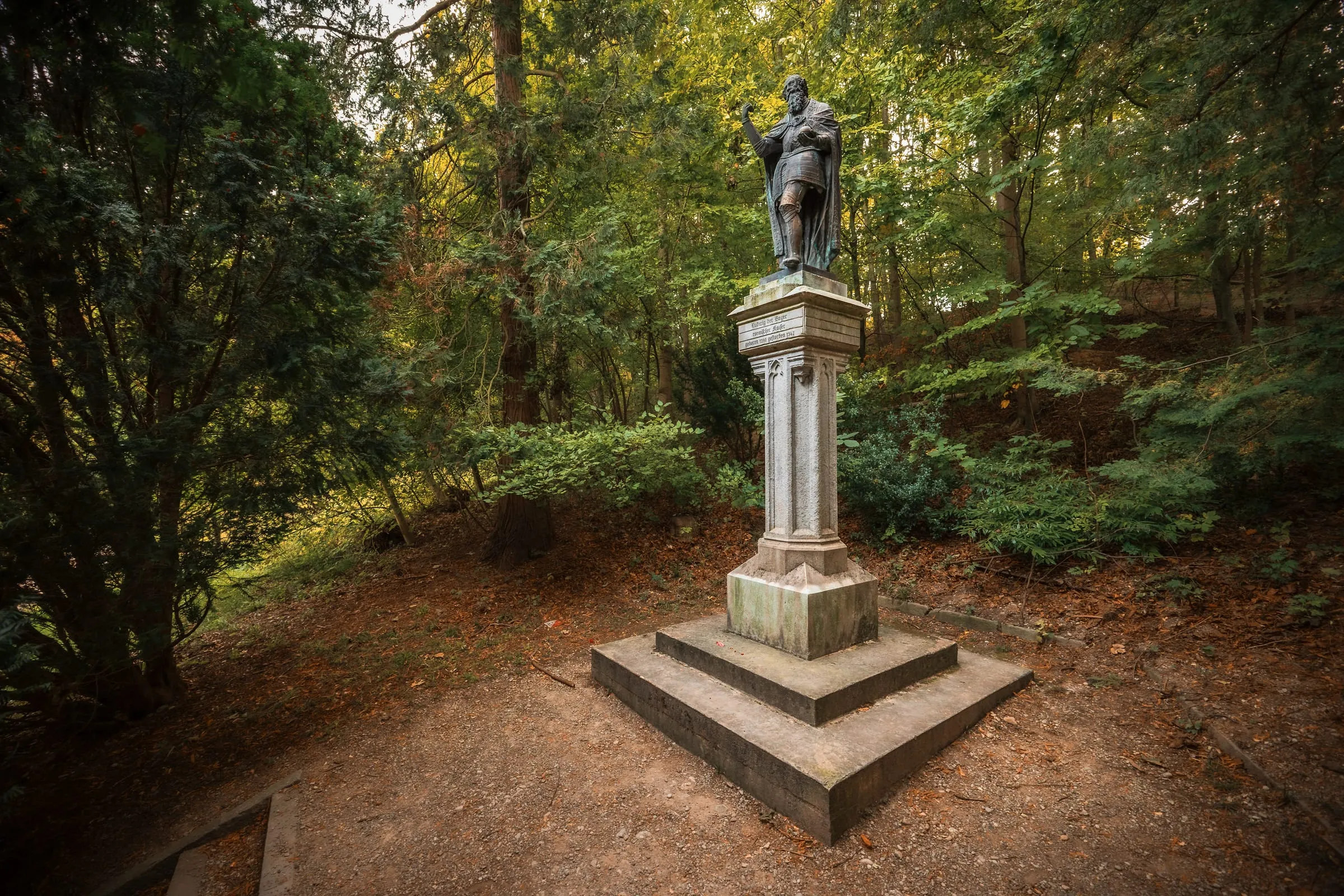 Bronzestatue Ludwigs des Bayern auf einem hohen Steinsockel mit Inschrift, auf einem laubbedeckten Platz zwischen grünen Bäumen im Hofgarten.