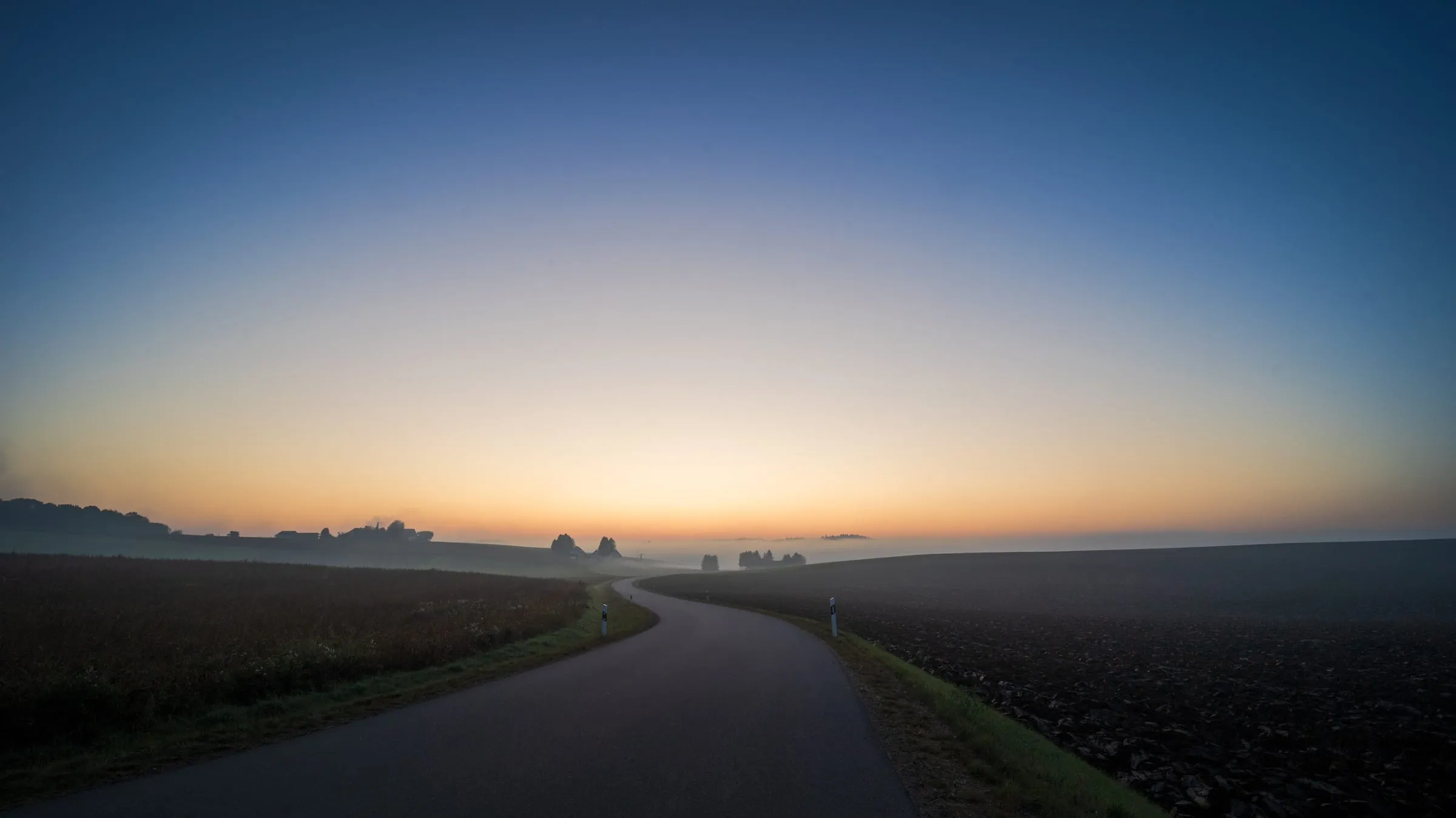 Eine schmale Landstraße führt in Kurven zwischen einem Feld und einem gepflügten Acker zum Horizont, der vor Sonnenaufgang orange leuchtet; in den Senken liegt Nebel.