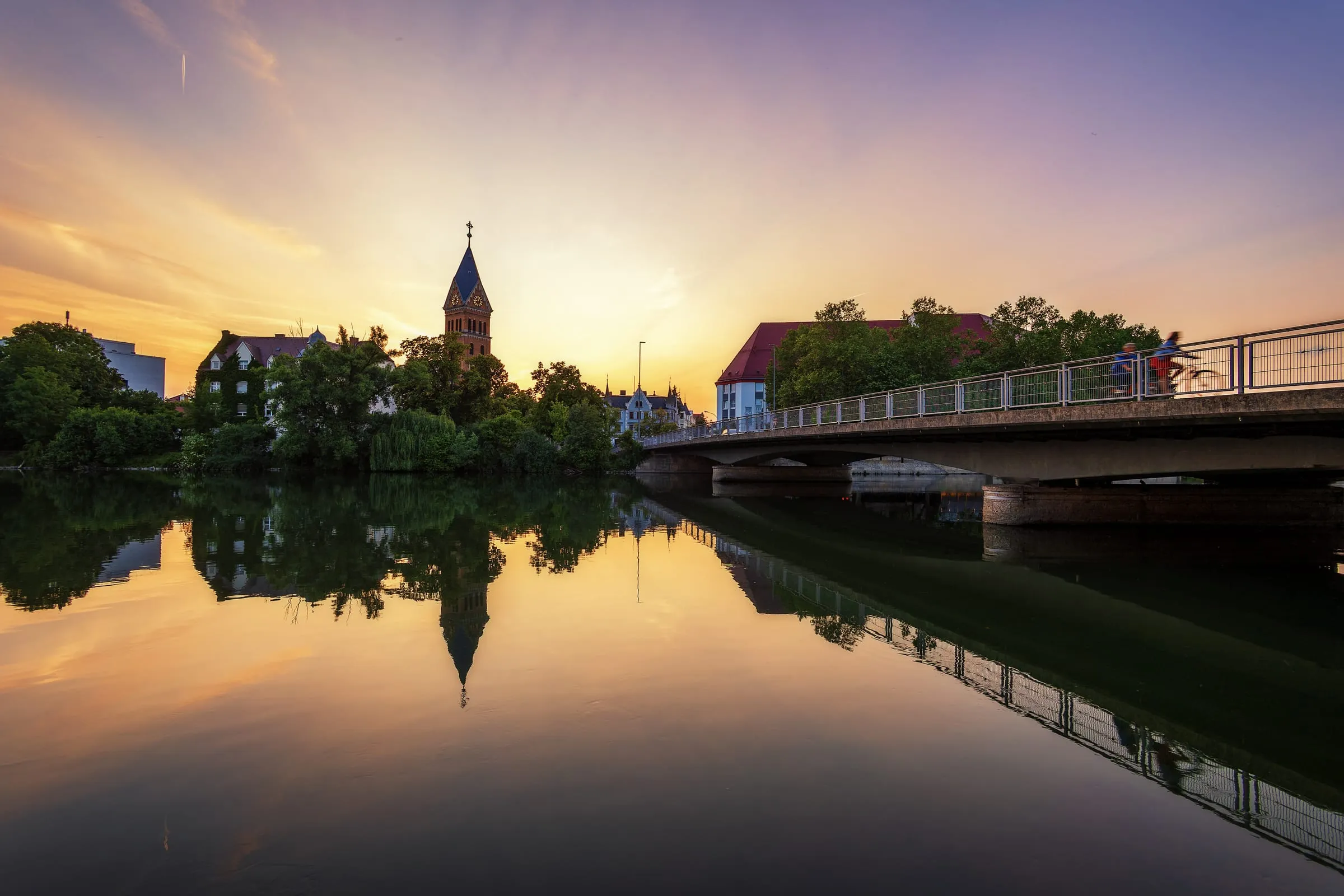 Die glatte Isar spiegelt Bäume, einen Kirchturm aus Backstein und eine Brücke mit zwei Radfahrern, darüber ein orange-violetter Himmel bei Sonnenuntergang.