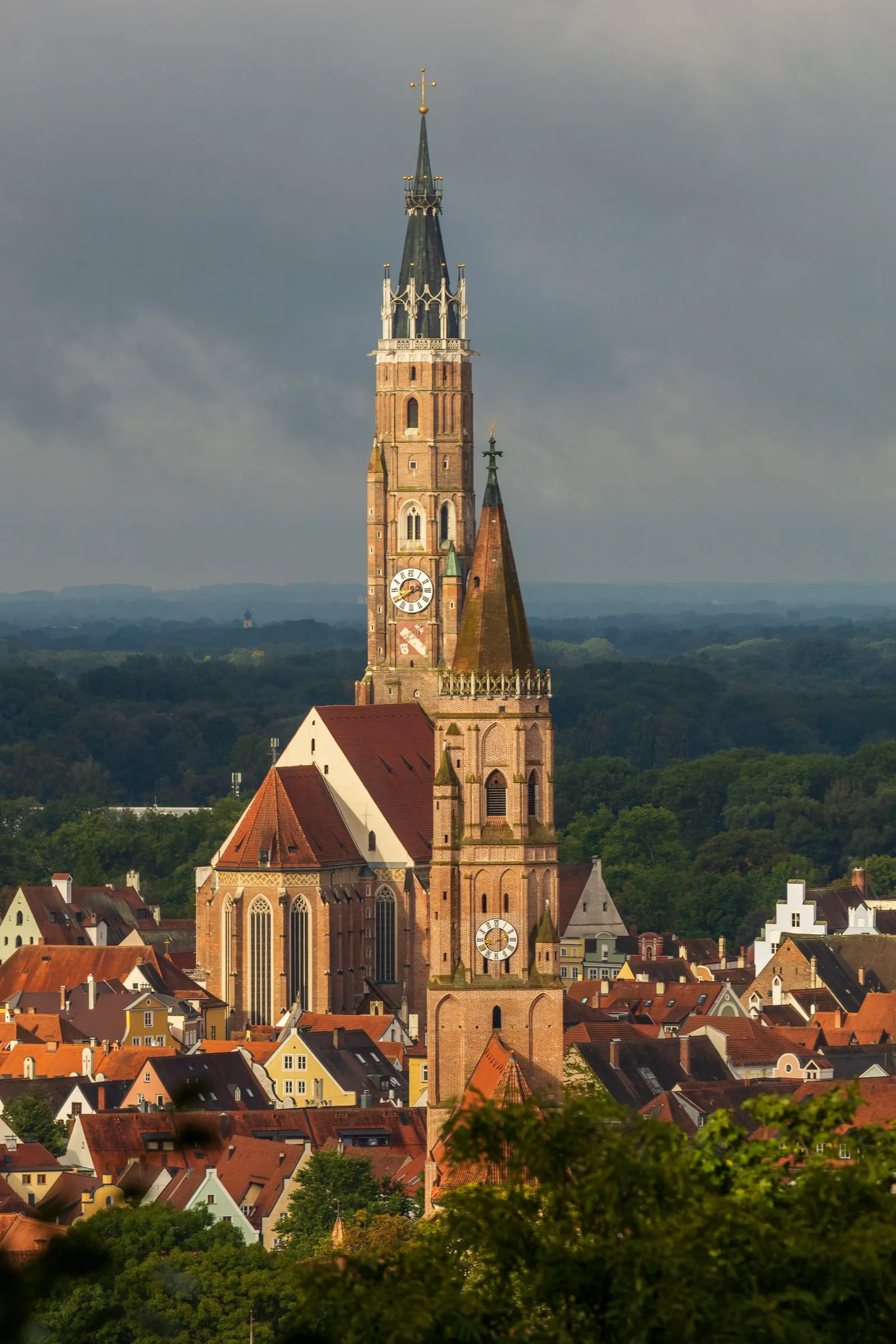 Der Backsteinturm von St. Martin ragt im warmen Morgenlicht vor dunklen Wolken auf, davor ein zweiter Turm, rote Altstadtdächer und dahinter Wald.